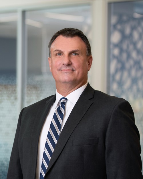 A portait of Steve Butz wearing a black suit jacket, white shirt, and a navy, white and silver striped tie.