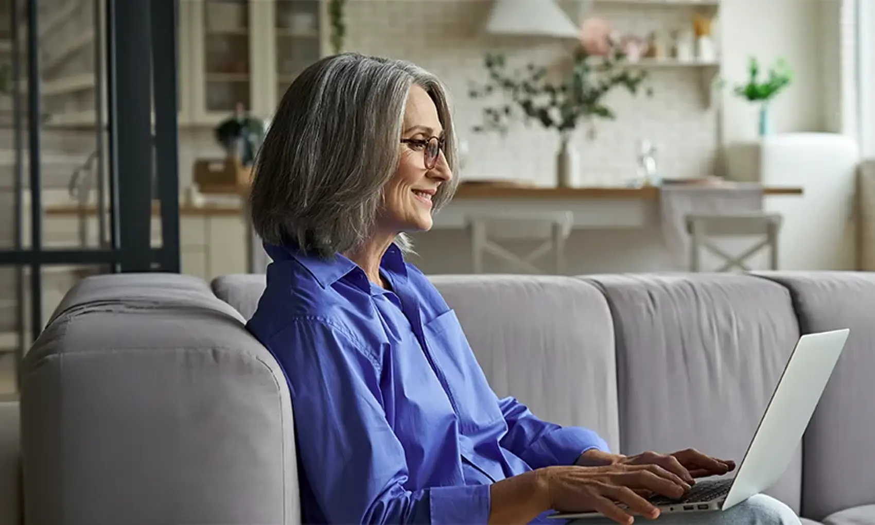 Happy 60s older mature middle aged adult woman holding laptop computer sitting on couch at home. 