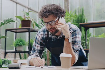 Image of a man wearing an apron talking on the phone and writing something down surrounded by a cup of coffee, a credit card processing machine, a laptop, and plants.