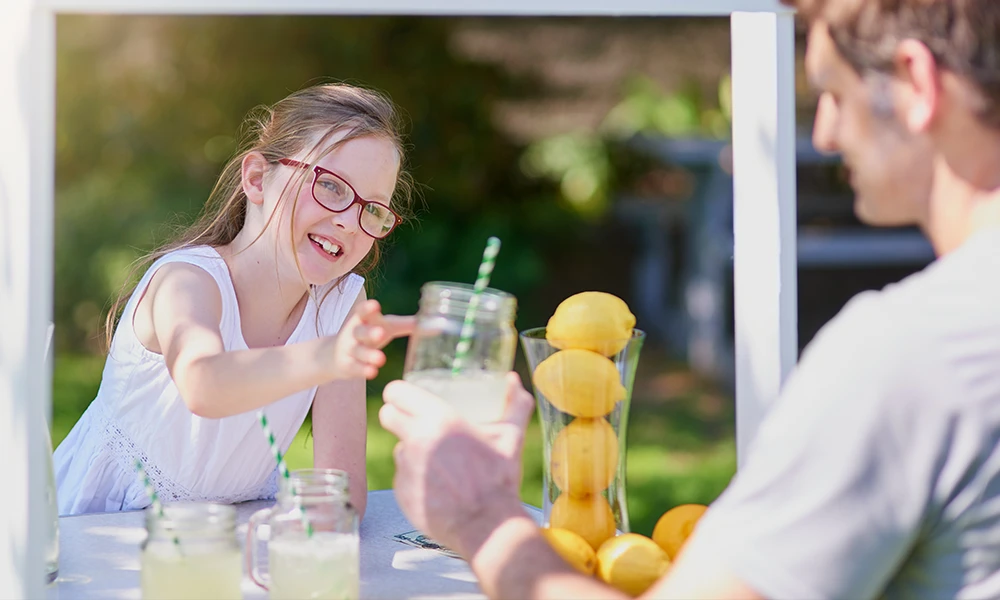 young girl hands glass of lemonade to a buyer at her stand