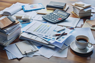 Image of a table covered in piles of documents, books, pens, a calculator and a cup of coffee.